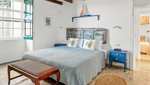 Une chambre avec un lit, un banc, une table de chevet, une étagère avec une maquette de voilier et une fenêtre avec une porte verte.
