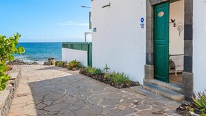 Un étroit sentier pavé de pierres mène à un bâtiment blanc doté d’une porte verte, un panneau indique une distance de 16 mètres, et on peut admirer la mer.