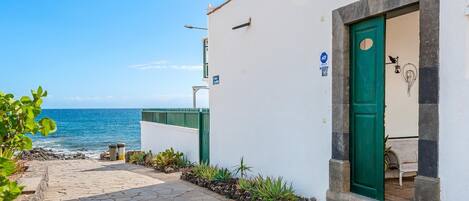 A narrow stone-paved path leading to a white building with a green door, a sign indicating a 16-meter distance, and a view of the sea.