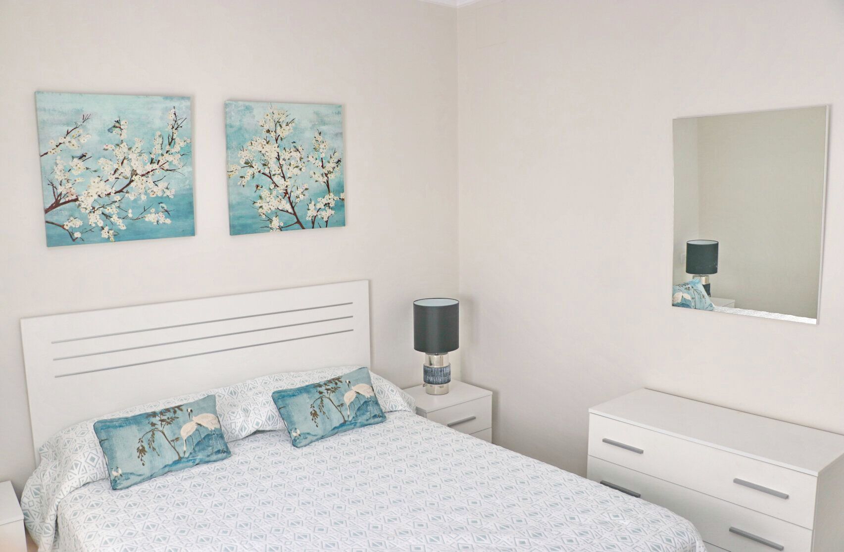 A bedroom with a bed, two wall-mounted paintings, a bedside lamp, a mirror, and a white dresser.