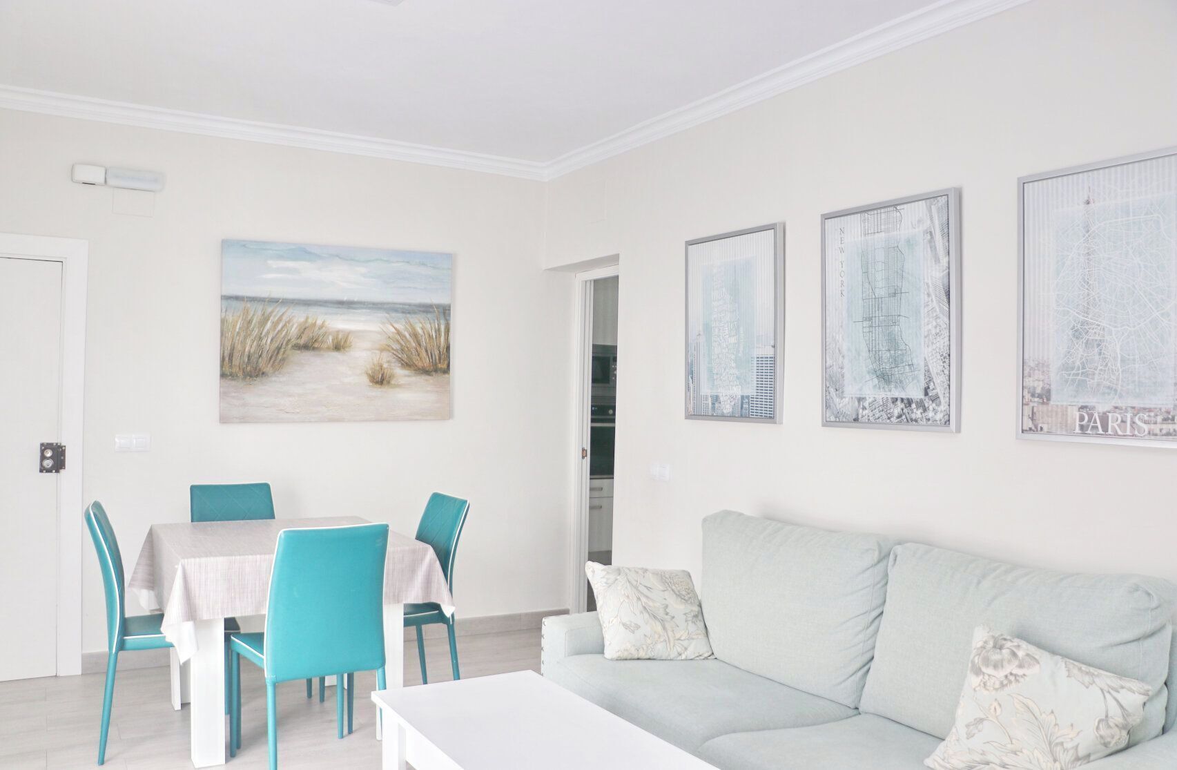 Una habitación con una mesa de comedor, sillas color turquesa, un sofá gris claro y tres cuadros con vistas urbanas en la pared.