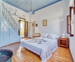 A bedroom with a wooden floor, a bed with a white lace cover, a nightstand, a hanging lamp, and a staircase with blue-painted railings. - Apartment with sea view (Simi)
