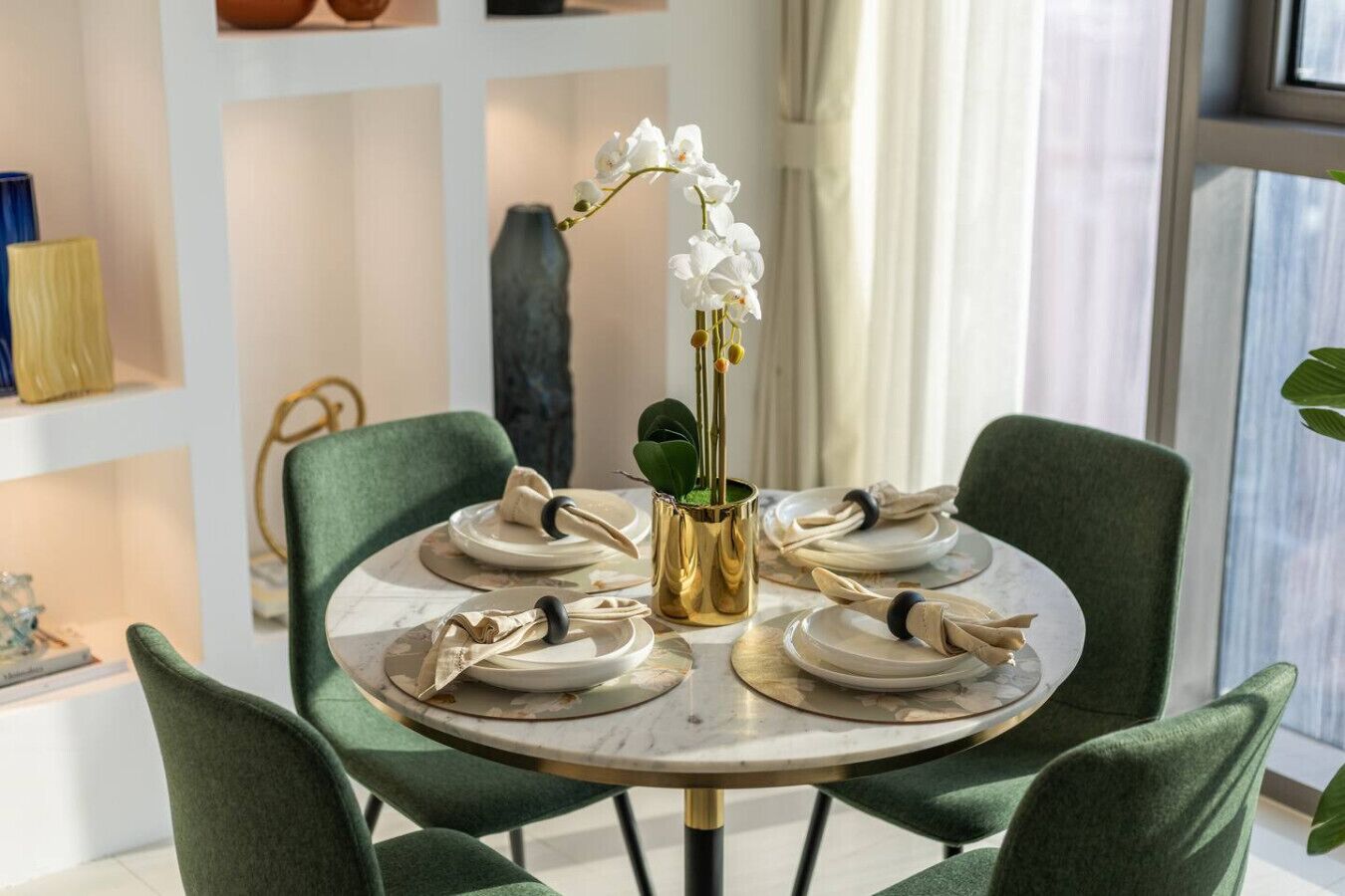 A round dining table with green upholstered chairs, white plates, and gold napkin holders.
