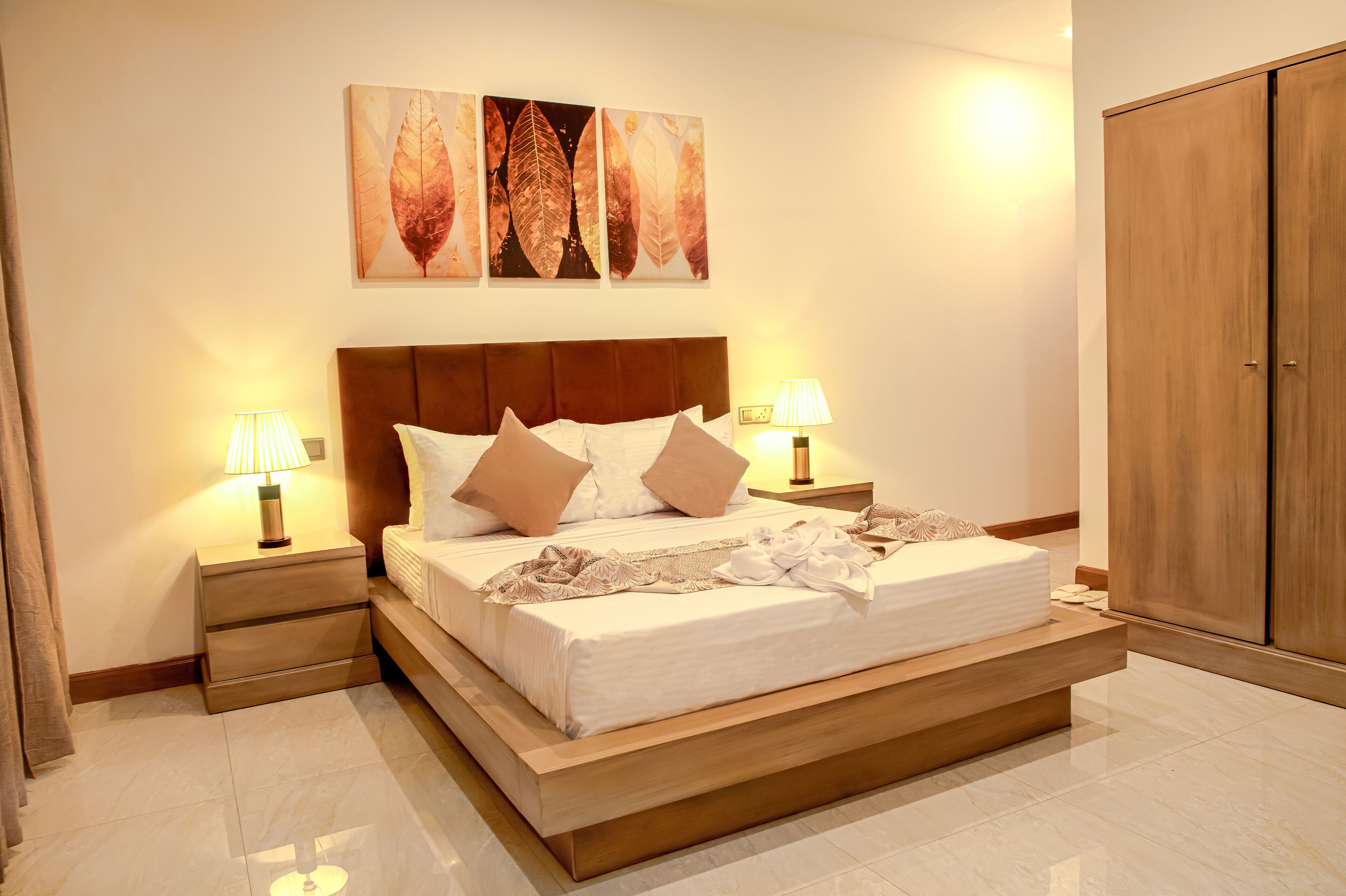 A modern bedroom with a large bed, two bedside tables with lamps, a wooden wardrobe, and three leaf-patterned wall art pieces.