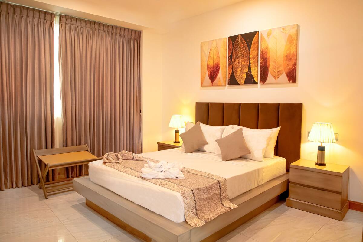 A hotel room with a large bed, two bedside tables with lamps, a bench, and a view of the outdoors through curtains.