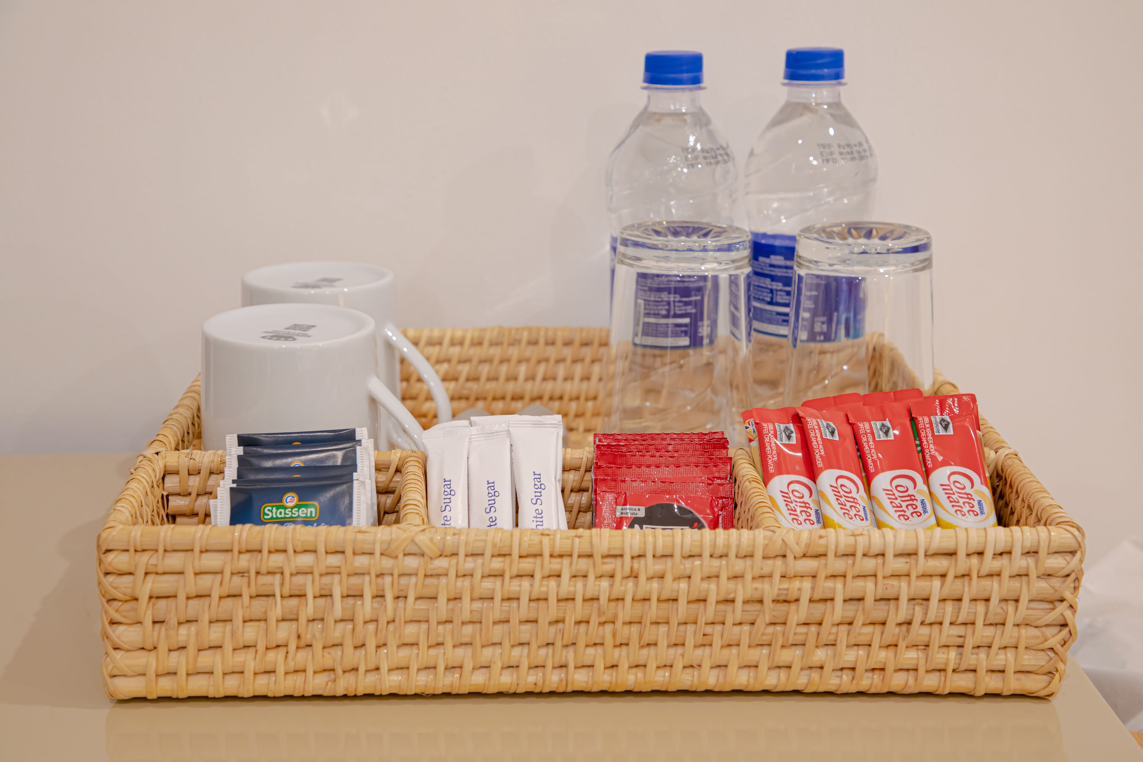 A wicker basket containing coffee packets, sugar packets, a white mug, and two water bottles.