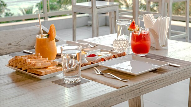 A table set with a plate of toast, a glass of water, a glass of juice, a glass of iced tea, and a glass of red drink with a straw and a slice of watermelon. - Ravo Hotel (Galle)