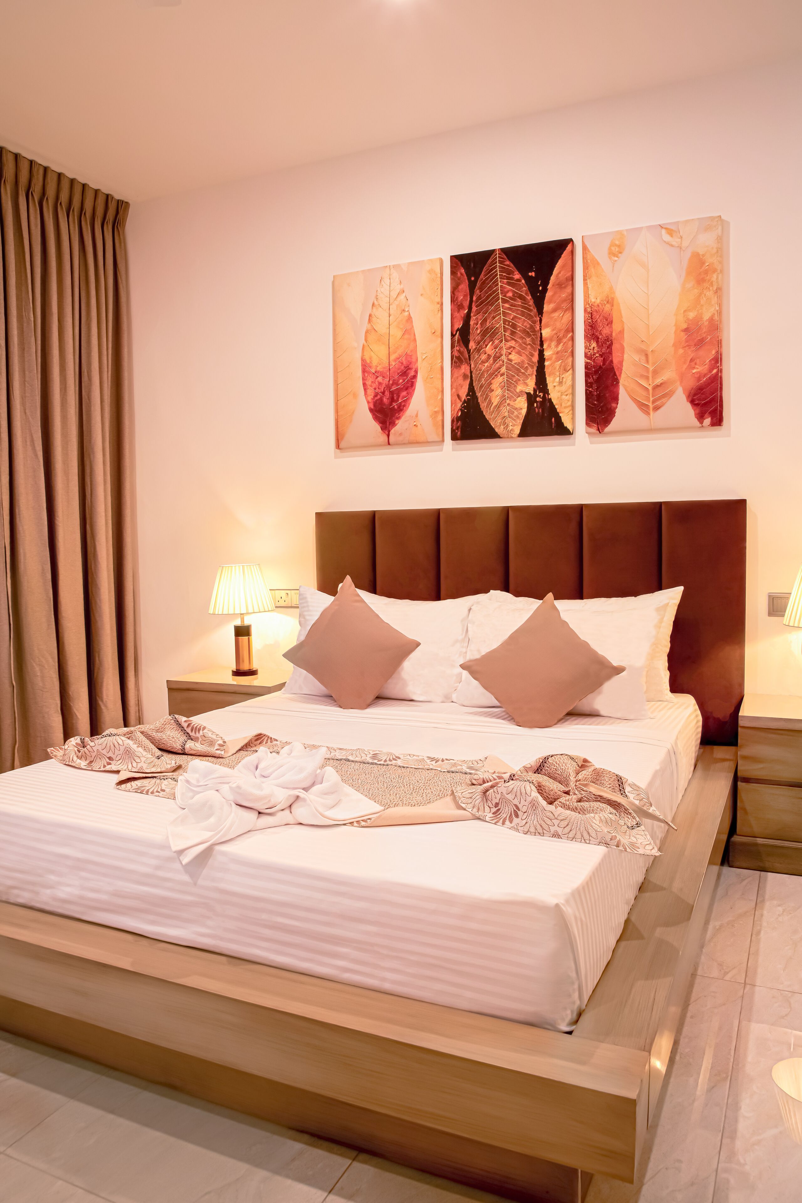 A neatly made bed with a brown headboard, a bedside table with a lamp, and a wall with three leaf-patterned paintings.