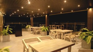 An outdoor seating area with wooden tables and chairs, potted plants, and a view of the city at night. - Ravo Hotel (Galle)