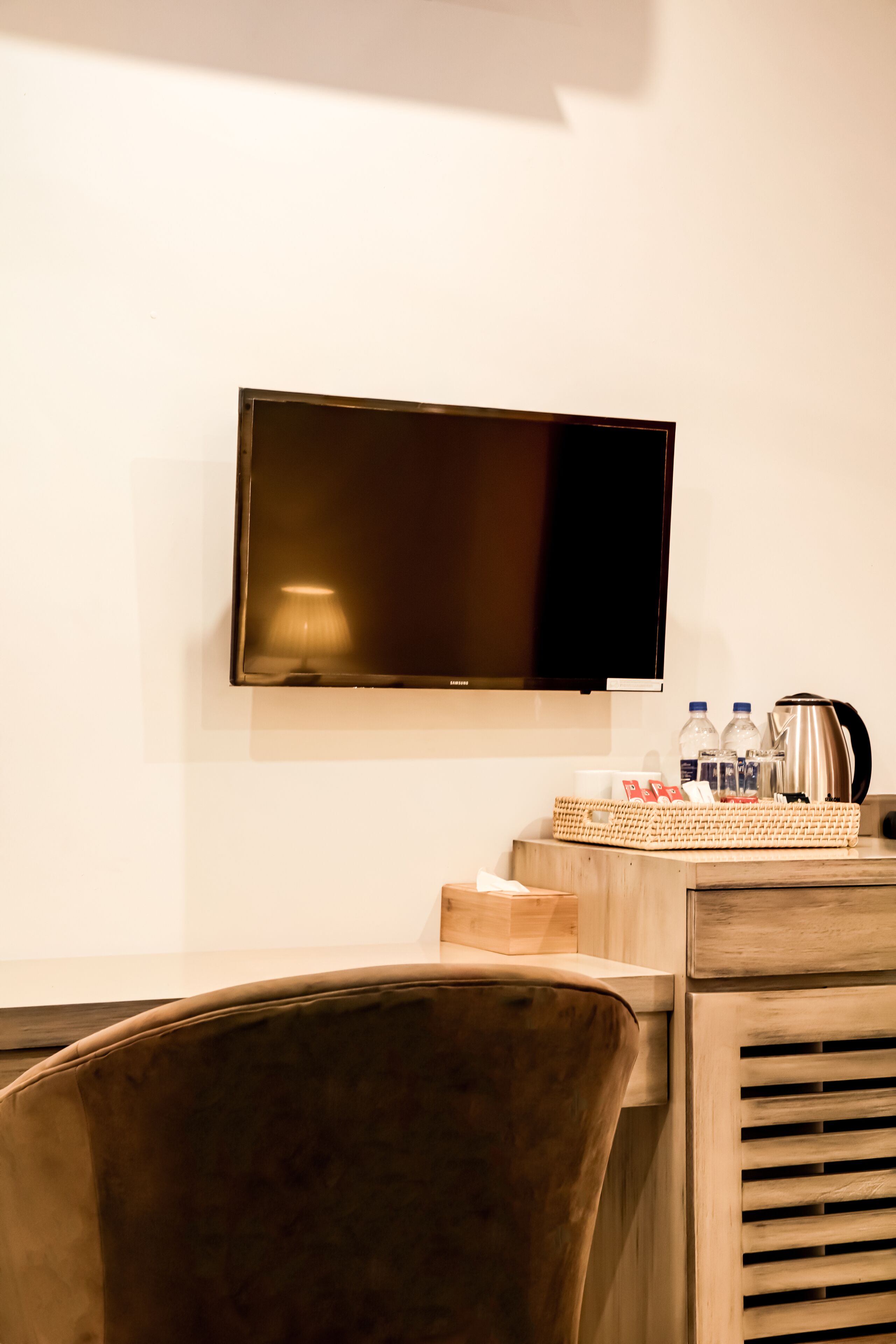 A hotel room with a flat-screen TV mounted on the wall, a wooden desk with a chair, and a basket containing toiletries.