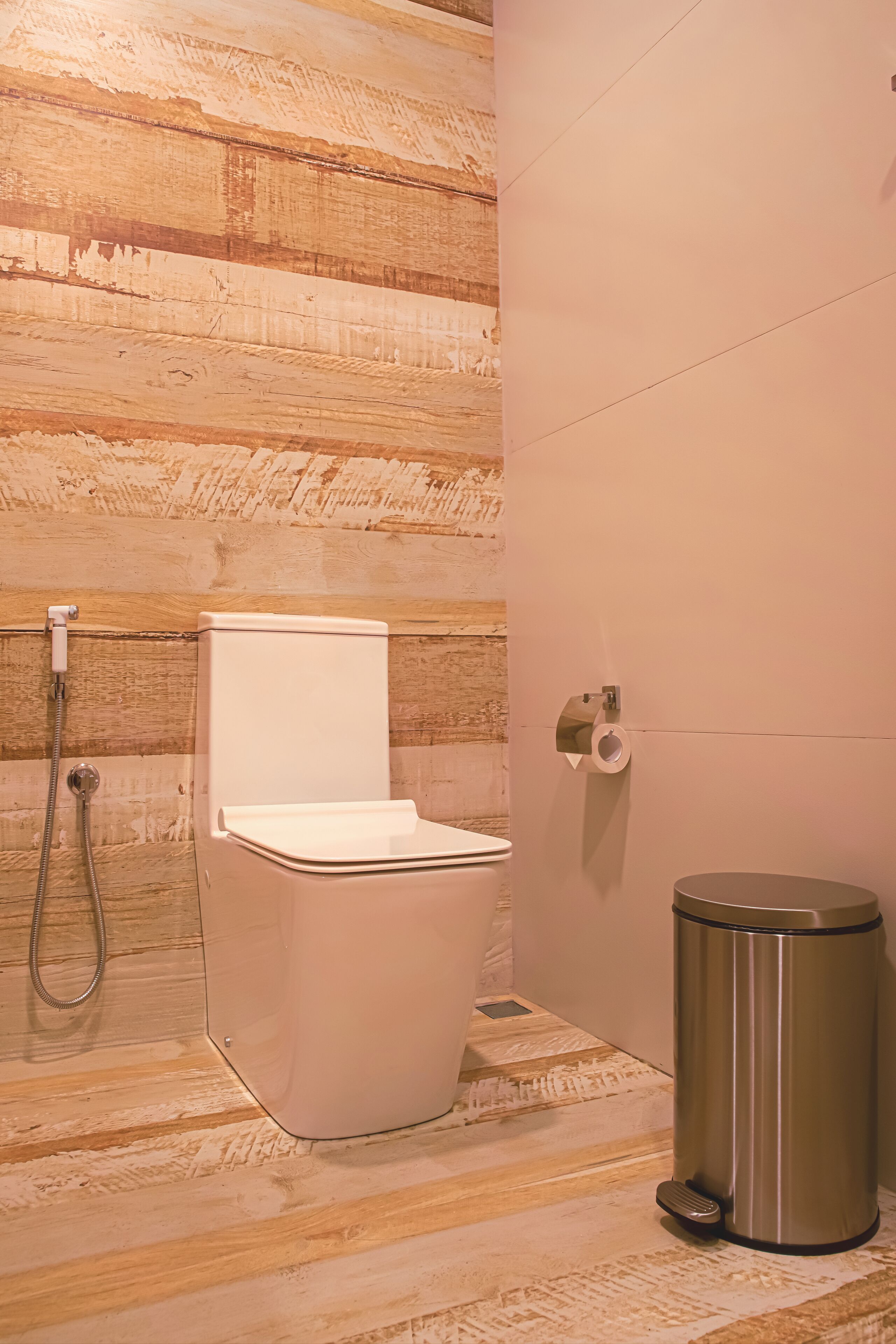 A bathroom with a wooden wall, a white toilet, a shower, and a trash bin.