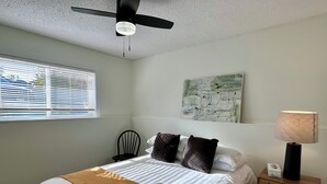 A neatly made bed with a white comforter and a mustard-colored throw blanket, a black chair, a wooden nightstand with a lamp, and a ceiling fan. - Beautiful Modern Two Bedroom Apartment  (Brandon)