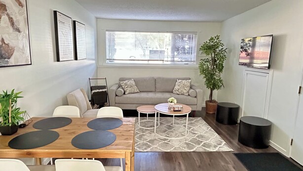 A modern living room with a wooden dining table, white chairs, a grey sofa, and a large window with blinds. - Beautiful Modern Two Bedroom Apartment  (Brandon)