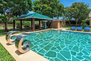 Uma piscina com um pavilhão de telhado verde, cadeiras de lounge e área gramada.
