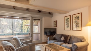 A cozy living room with a checkered sofa, a wooden coffee table, and a television. There are framed botanical prints on the wall and a window with a view of trees. - Casa Funivia, Casa Funivia, close to the center and the cableway (Cortina d´Ampezzo)