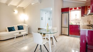 Uma sala de estar moderna com um sofá branco, uma mesa de jantar de vidro e cadeiras brancas. Há uma área de cozinha com armários vermelhos e uma geladeira de aço inoxidável.