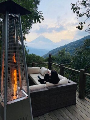 A person relaxing on a wicker sofa with a glass-top table, overlooking a mountainous landscape.