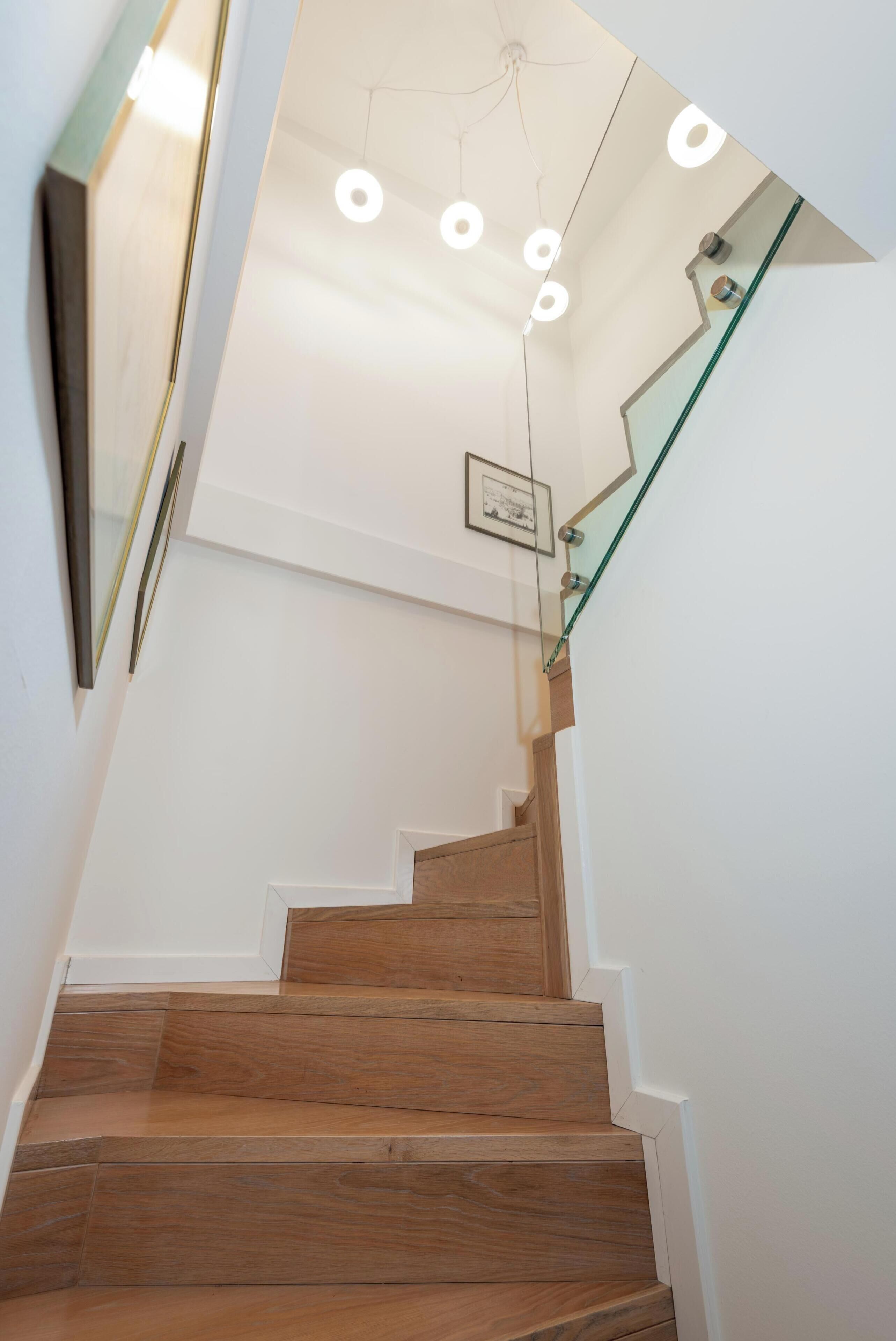 Una escalera con escalones de madera, barandilla de vidrio e iluminación circular en el techo.