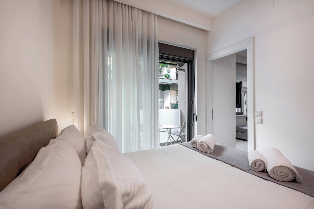 Quarto de hotel com uma cama grande, área de estar com mesa e cadeiras, e vista para a varanda externa.