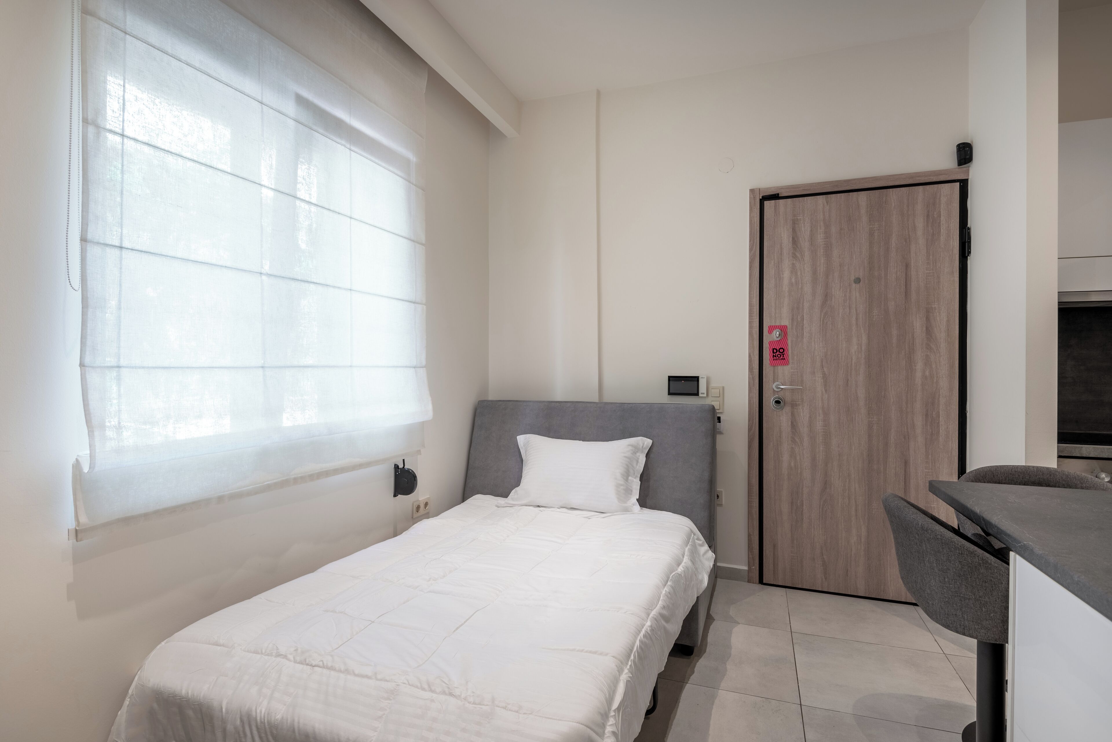 A bedroom with a bed, a window with blinds, a door, and a kitchenette area.
