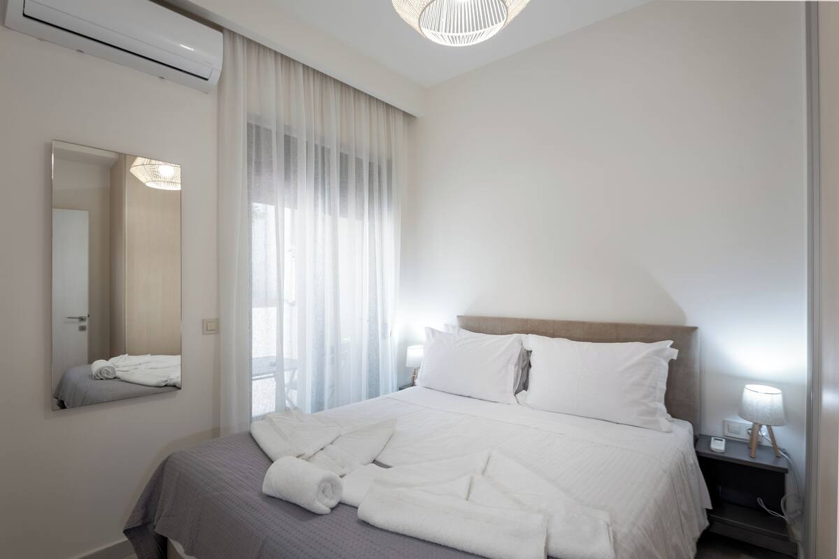 Quarto de hotel moderno com uma cama grande, mesa de cabeceira com abajur, espelho e janela com cortinas translúcidas.