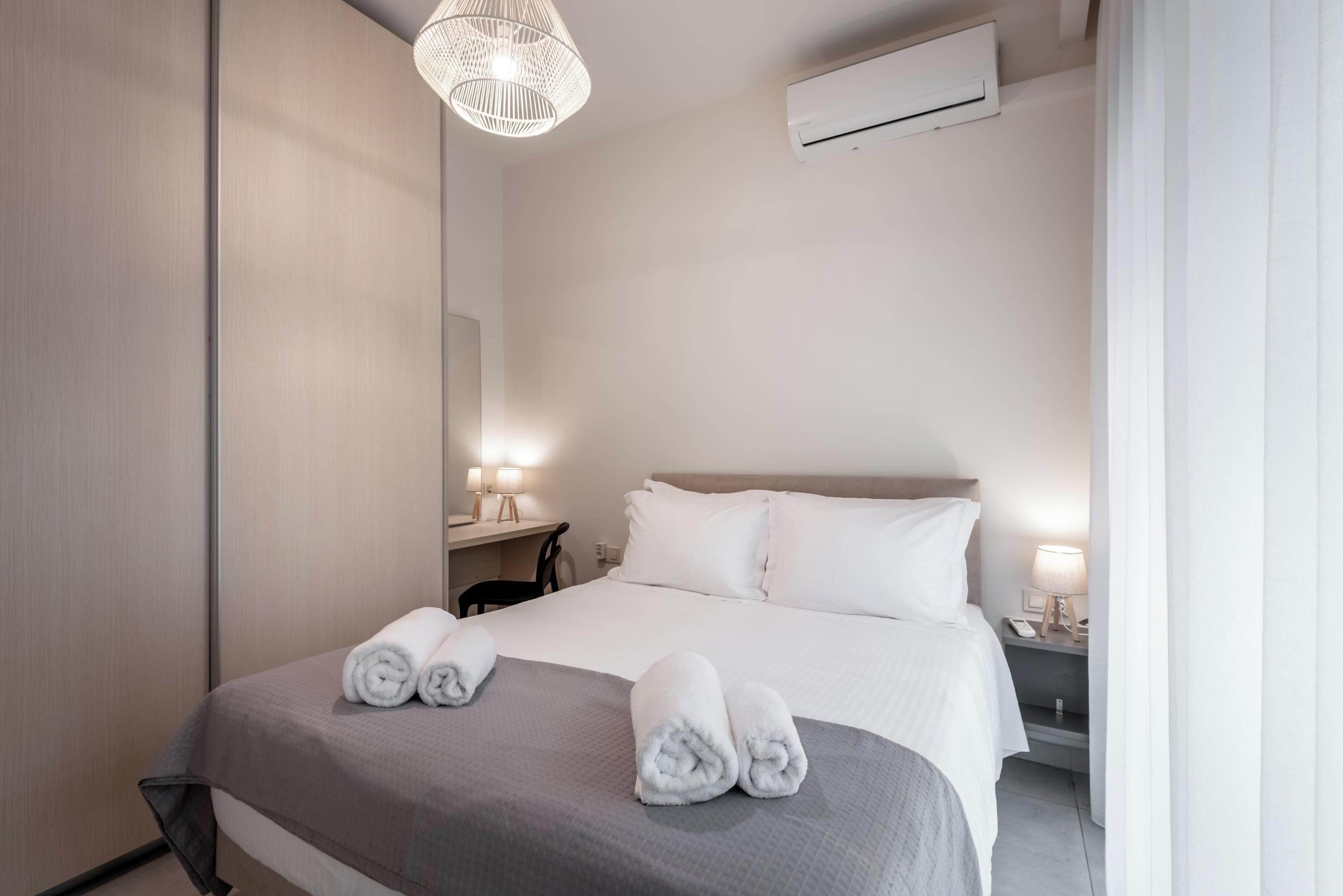 A hotel room with a bed, bedside tables, a desk, a chair, and a hanging light fixture.