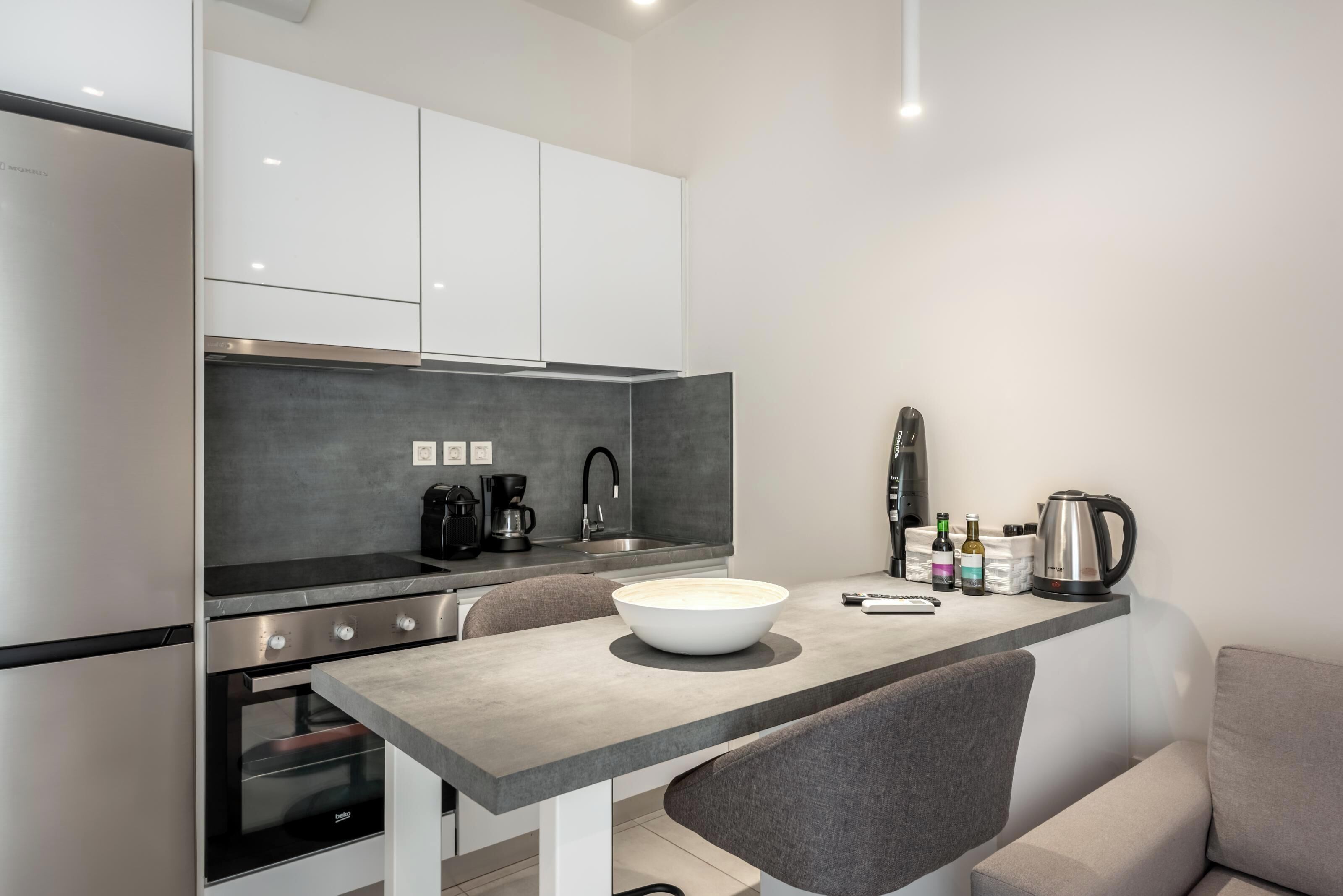 Uma cozinha moderna com armários brancos, bancada cinza e forno embutido. Há uma tigela branca grande na bancada e uma chaleira.