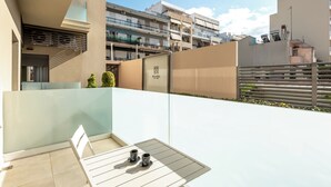 A modern outdoor seating area with a table and chairs, a glass partition, and a view of surrounding buildings. - Harma City Living (Chania)