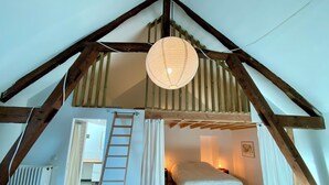 A loft bedroom with a sloped ceiling, wooden beams, a ladder, a bed, and a hanging light fixture. - Spacious houses in Normandy, aux Alpes Mancelles (Moulins-le-Carbonnel)