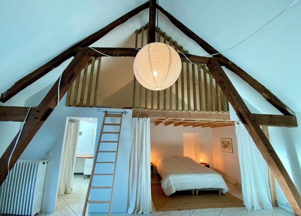 A loft bedroom with a sloped ceiling, wooden beams, a ladder, a bed, and a hanging light fixture.