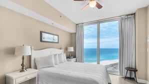 Un dormitorio con una cama grande, dos mesitas de noche, un ventilador de techo, vista a la playa y un cuadro en la pared.