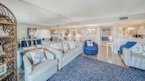 A spacious living room with a large sofa, a blue armchair, and a dining area with a kitchen in the background.