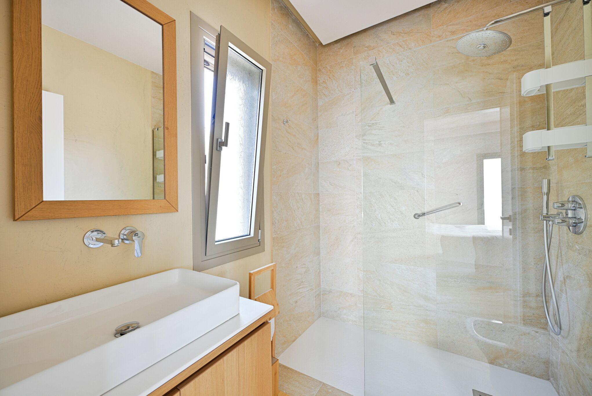 A modern bathroom with a large mirror, a white sink, a shower with a glass door, and a window providing natural light.