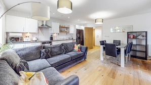 A modern living room with a grey sofa, a dining table with chairs, and a kitchen area with white cabinets and a black bookshelf. - Modern 2Bed - 5min to Highbury & Islington Station (London)