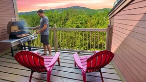Un homme prépare un barbecue sur une terrasse extérieure, avec une vue sur une chaîne de montagnes.