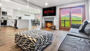 Un salon moderne avec une cheminée, un canapé, un pouf à motifs et un coin cuisine visible au fond.
