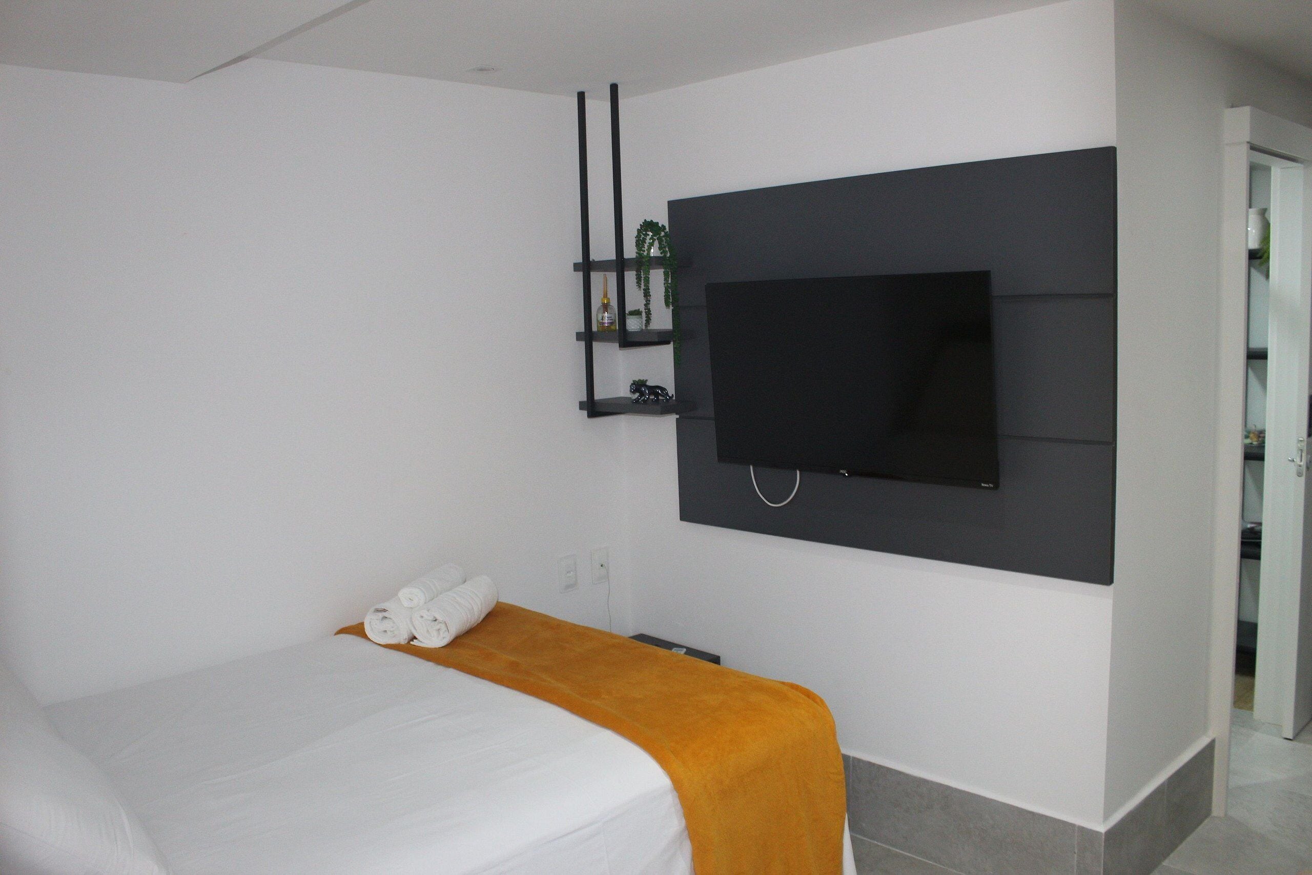 A modern hotel room with a bed, a TV mounted on the wall, and a small shelf with decorative items.