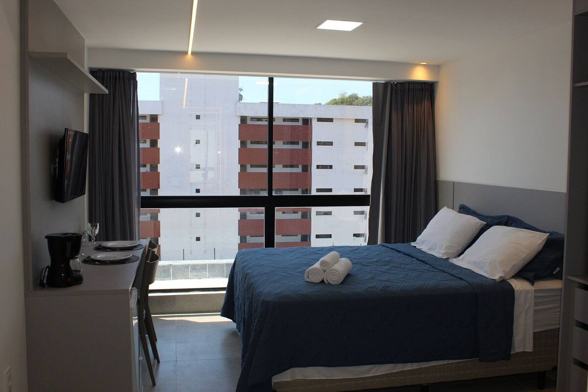 Habitación de hotel con cama, televisión, zona de comedor con mesa y sillas, y vistas a edificios desde la ventana.