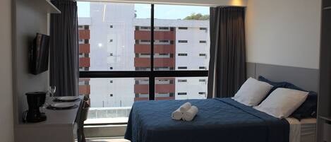 A hotel room with a bed, a TV, a dining area with a table and chairs, and a view of buildings through the window.