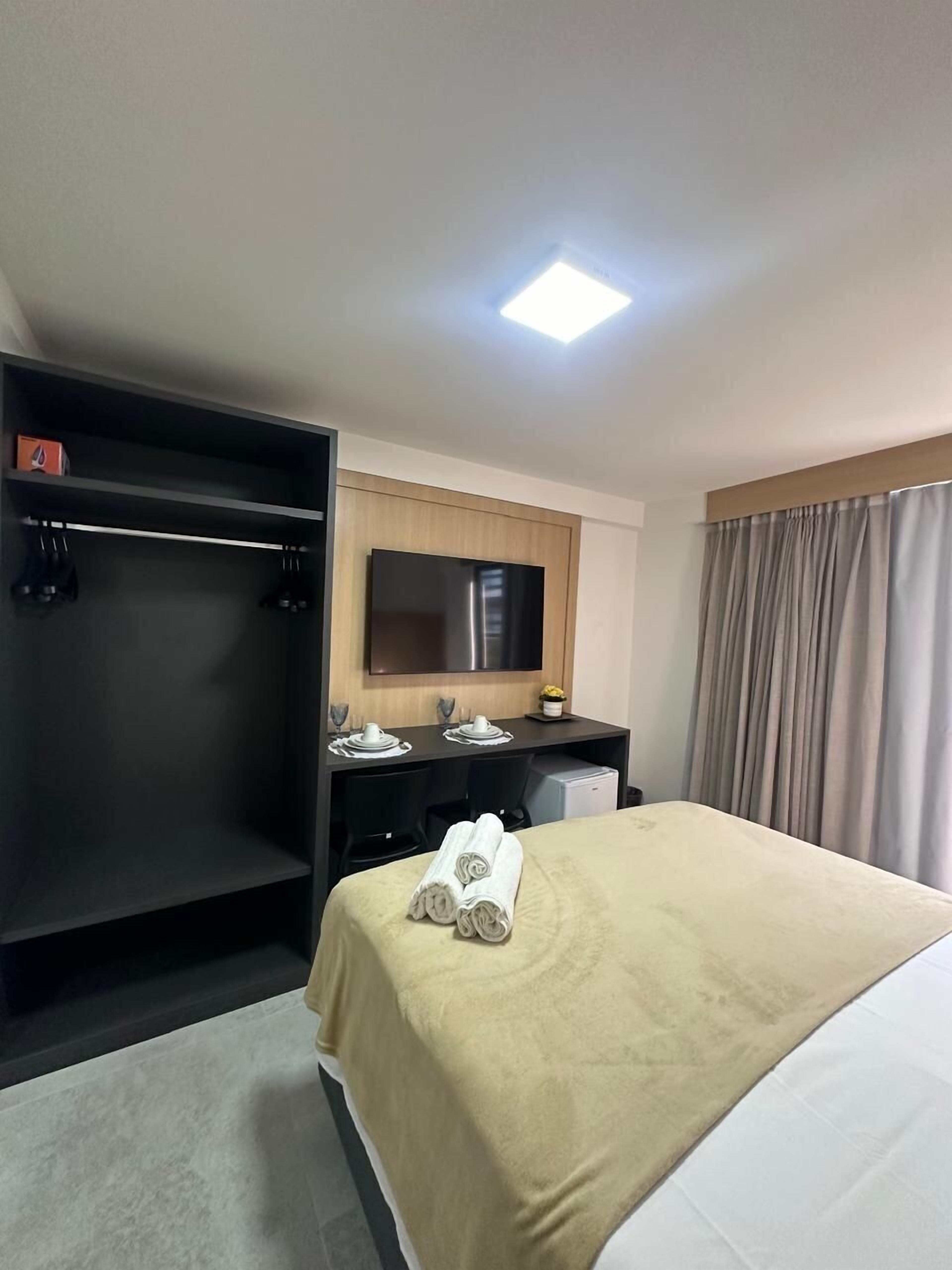 Quarto de hotel com uma cama, escrivaninha, cadeira, televisão e guarda-roupa.