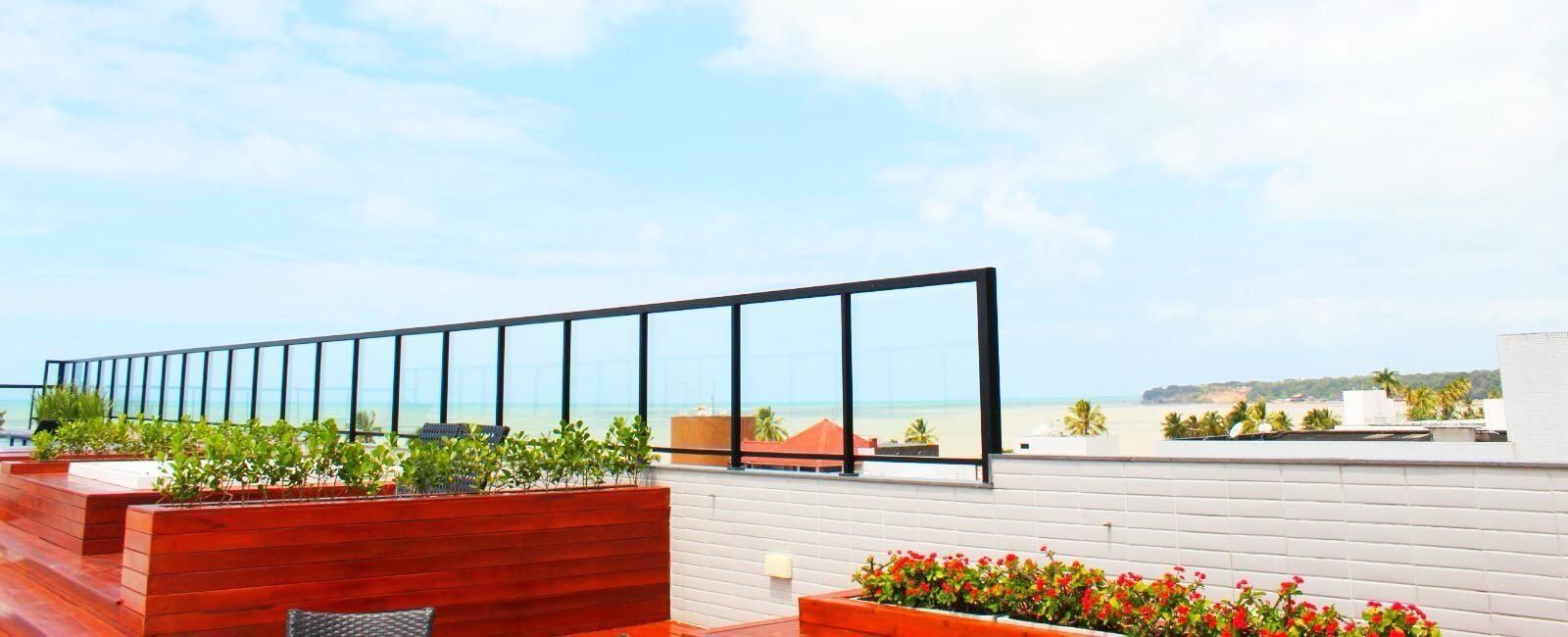 A rooftop terrace with a glass balustrade, wooden planters with greenery, and a view of the beach.