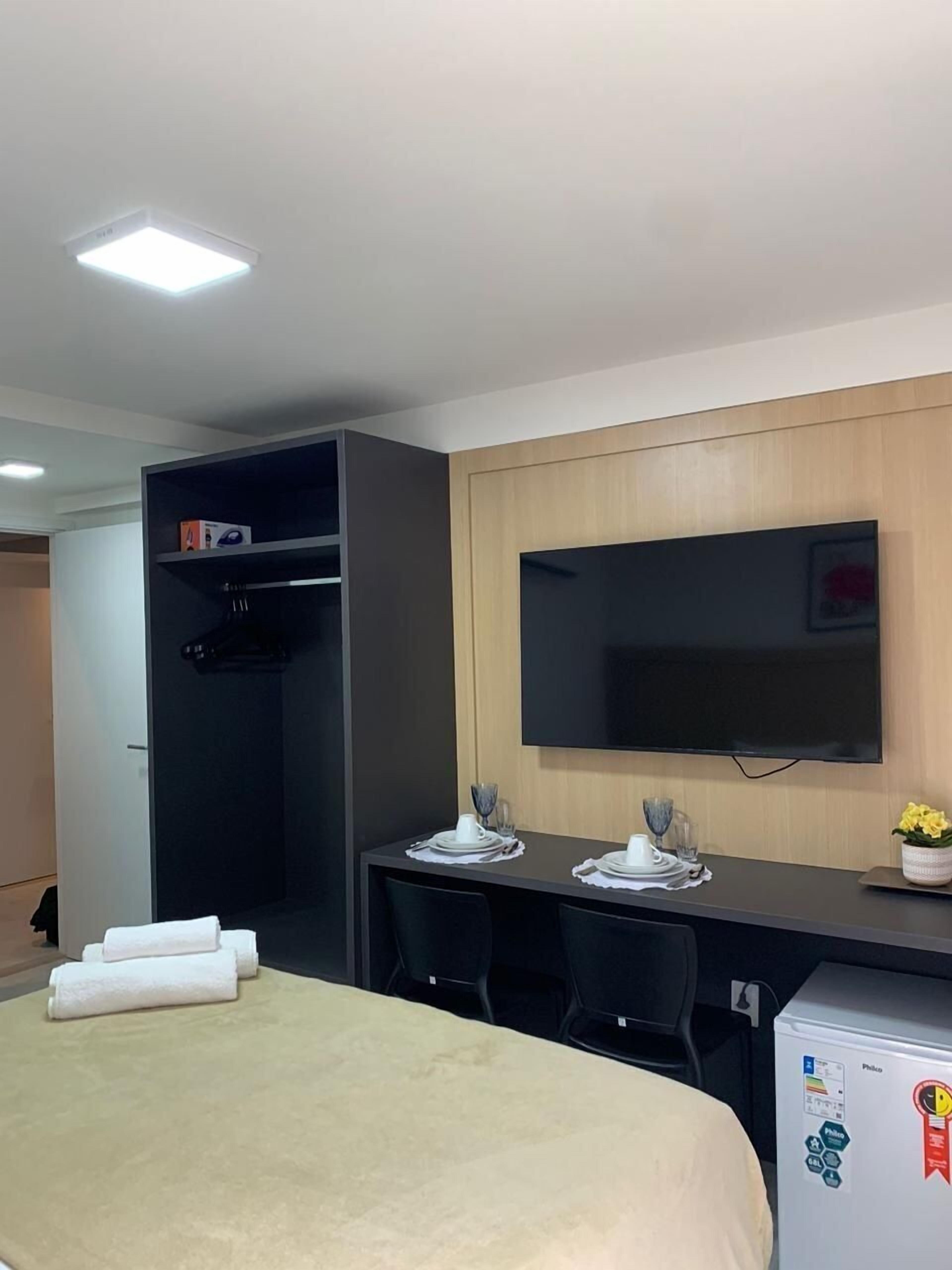 Habitación de hotel con cama, escritorio, silla, televisor montado en la pared y armario.