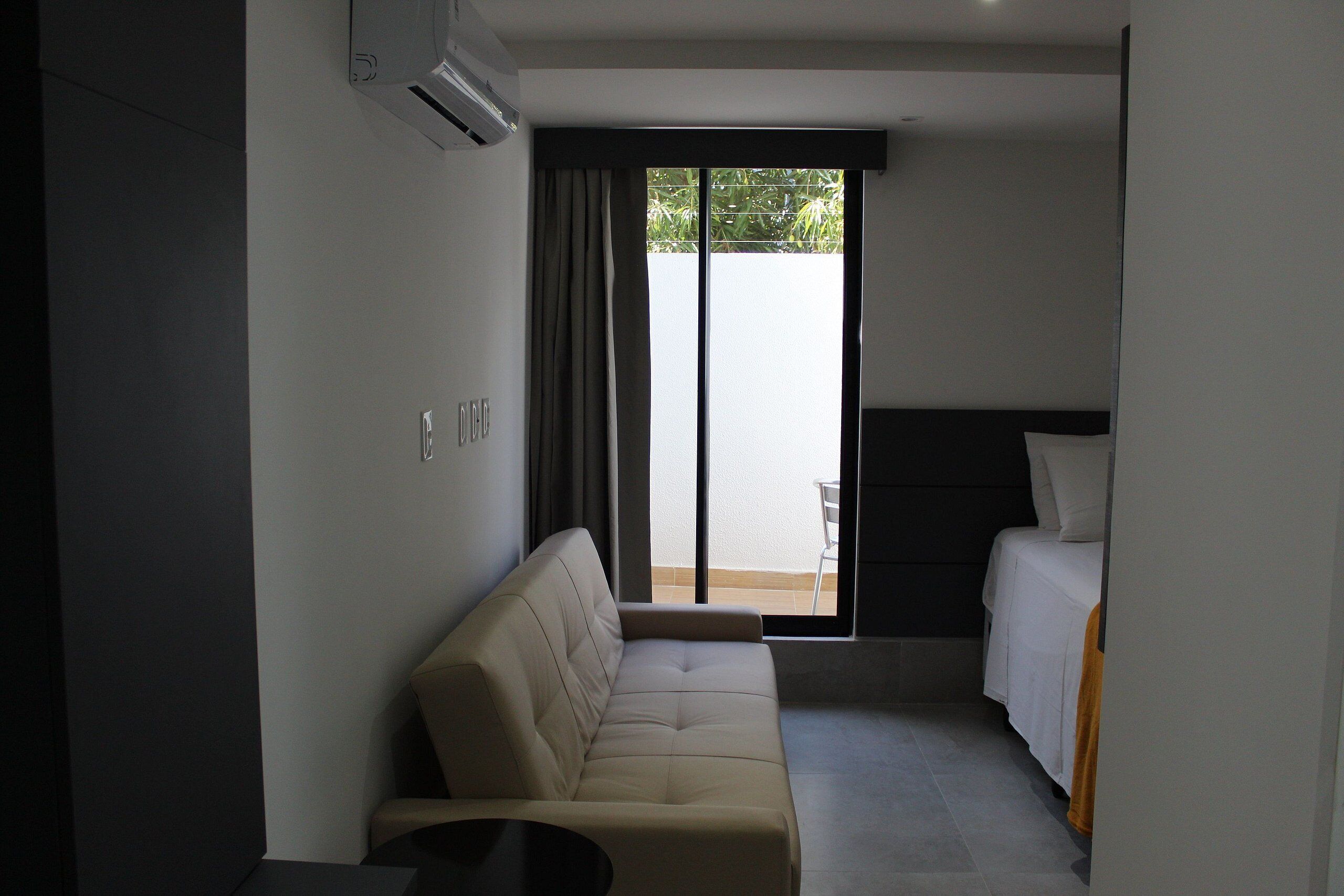 Una habitación de hotel moderna con sofá, cama, una mesita y un ventanal con cortinas.