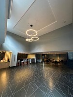Um lobby espaçoso com design moderno, destacando um balcão de recepção, um bar e uma área de refeições com várias mesas e cadeiras.