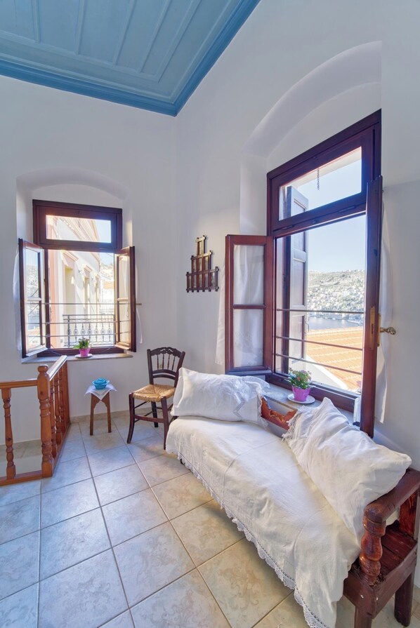 A cozy room with a bed, a chair, a small table, and two windows offering a view of the city.