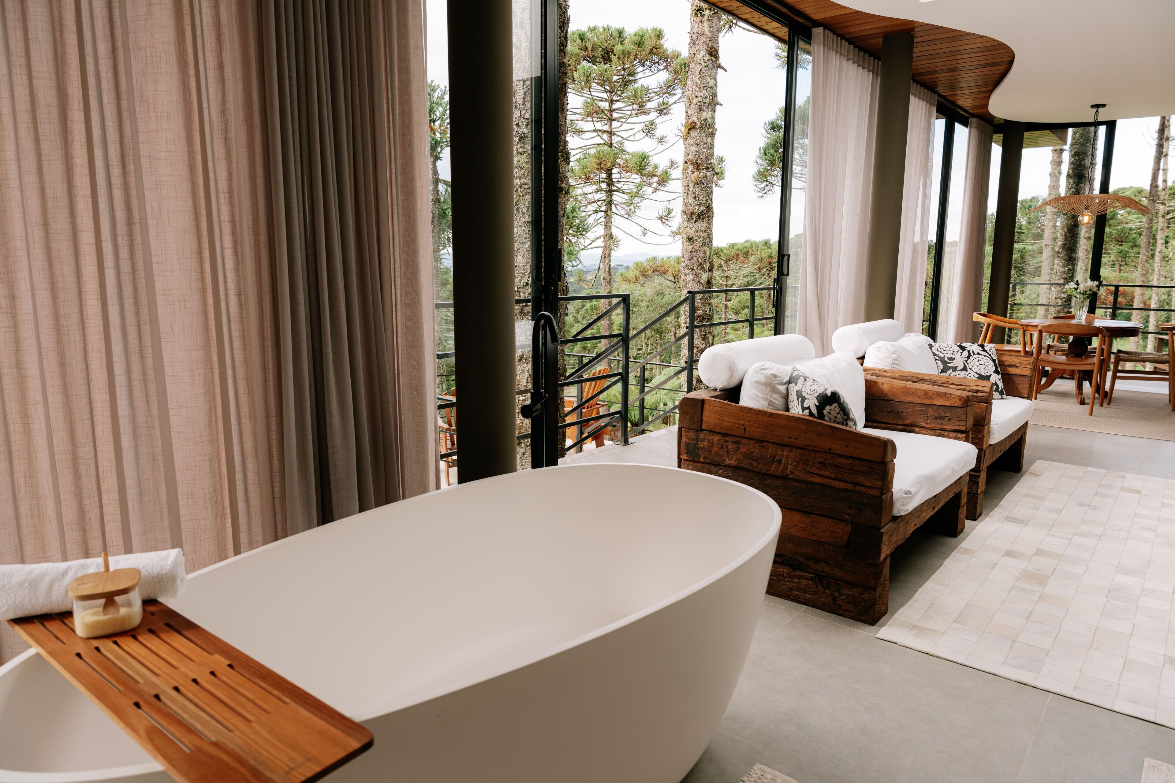 A modern bathroom with a freestanding bathtub, a wooden bench with cushions, and a large window offering a view of trees.