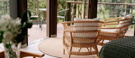 A wicker chair with a cushion, a round woven mat, and a wooden table in a room with large windows.