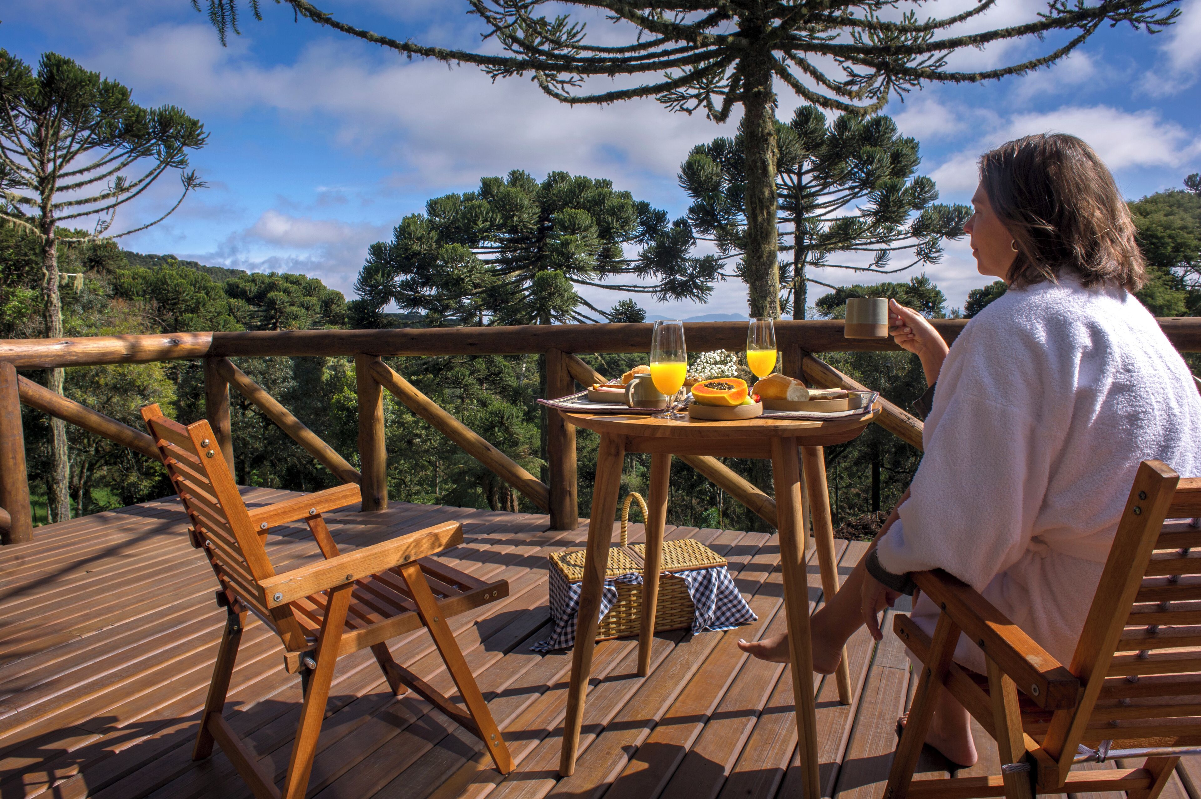 A wooden deck with a table set for two, a wicker basket, and a view of trees and hills.