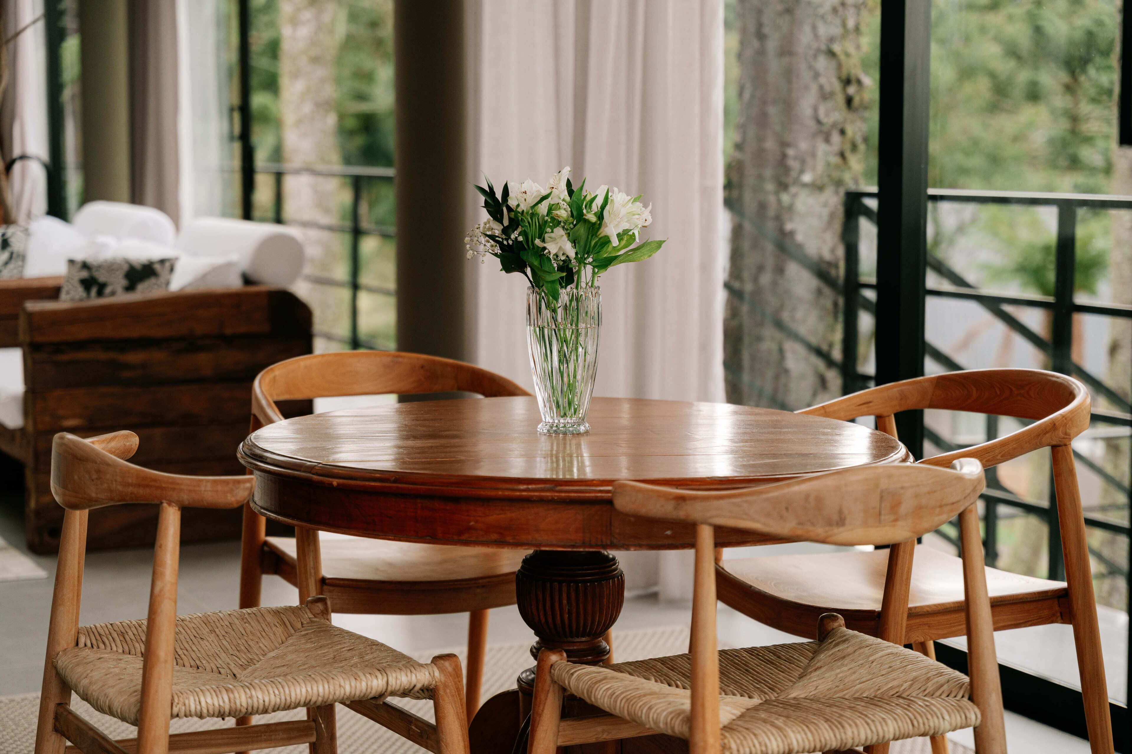 Een ronde houten tafel met een glazen vaas gevuld met witte bloemen, omringd door vier houten stoelen met geweven zittingen.