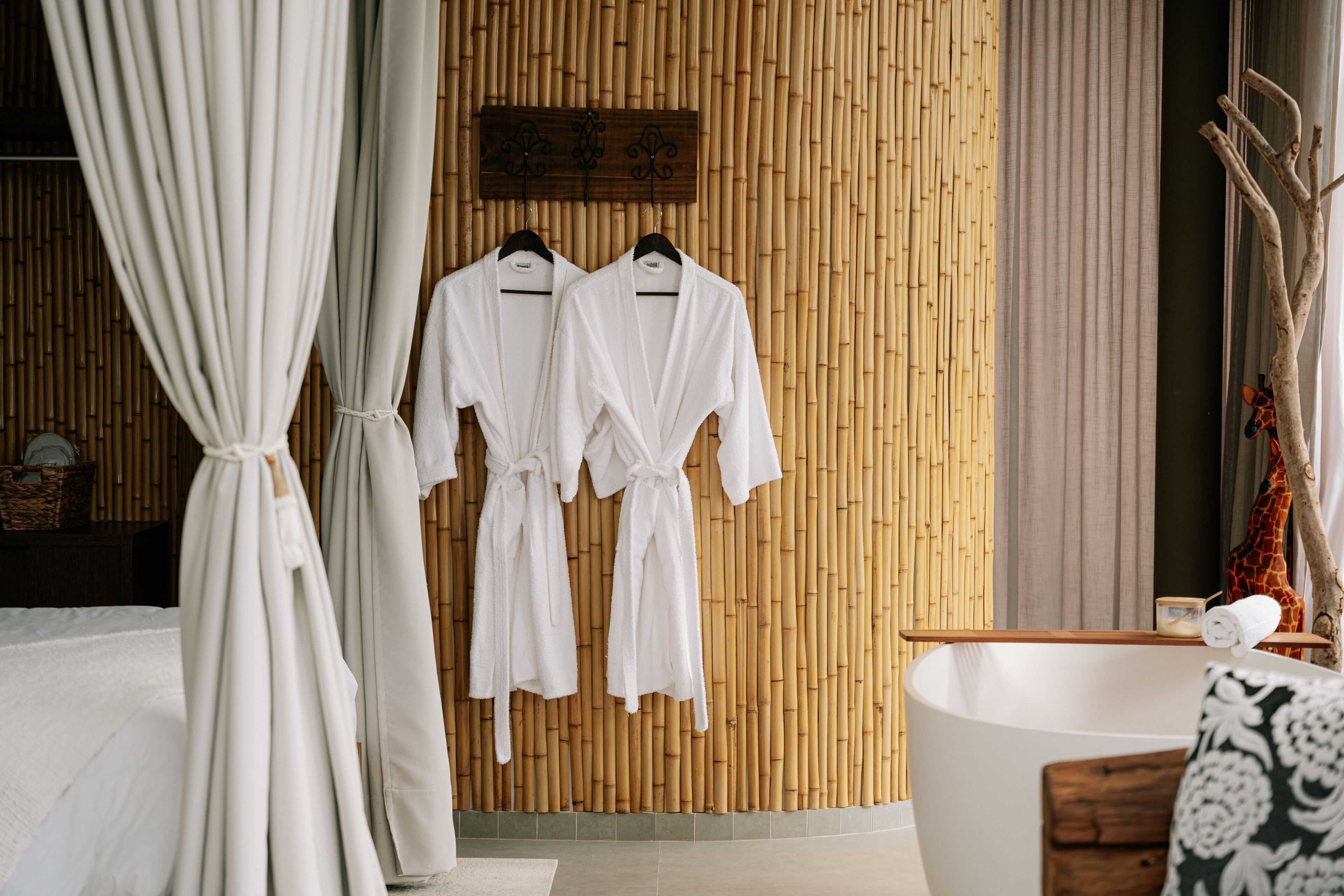 A room with a bamboo wall, a white bathtub, and two white robes hanging on a rack.